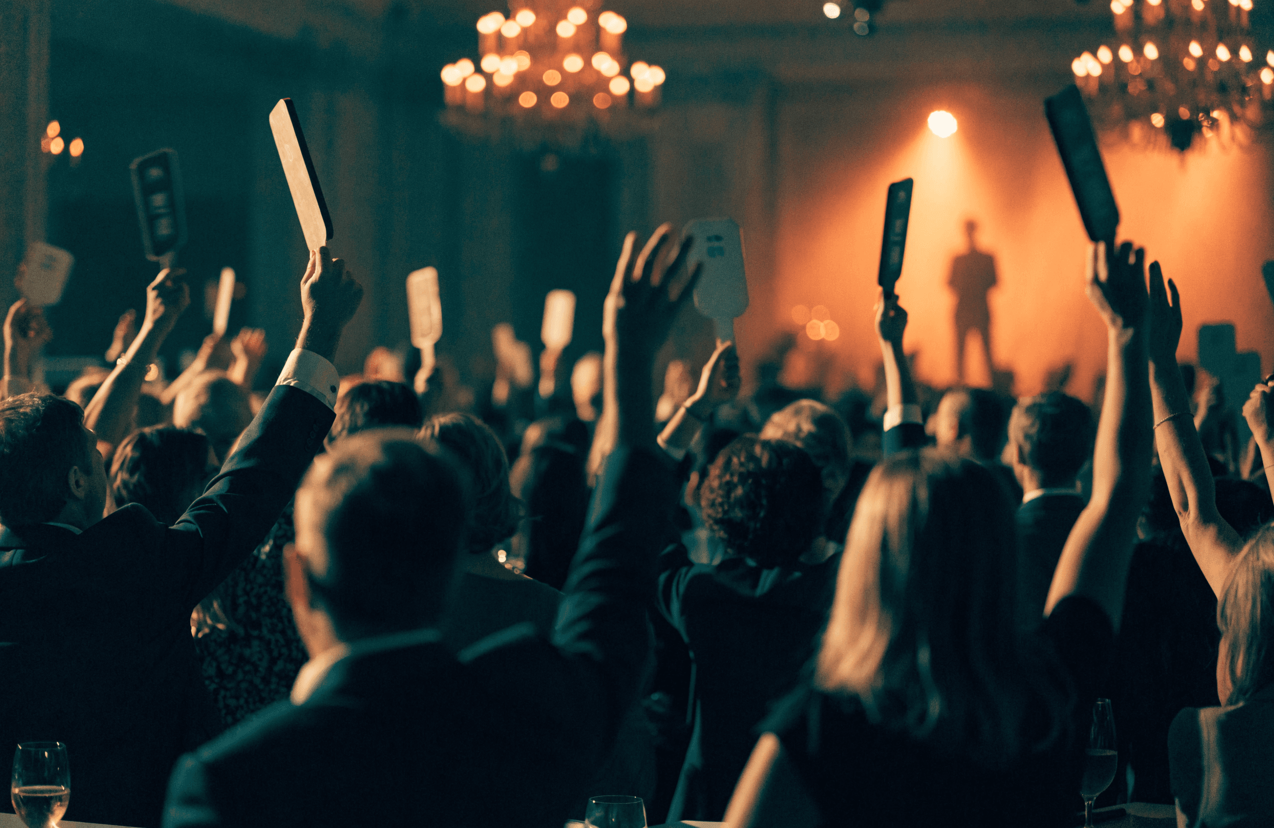 A warm ballroom at the peak of a fundraiser ask, a sea of raised bidding paddles under chandelier light